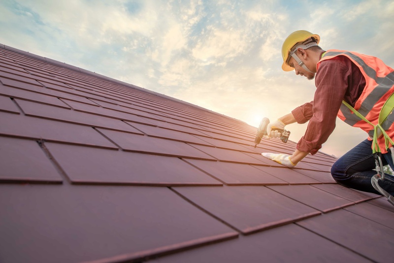 Roofer Working In Special Protective Work Wear Gloves Using Air Or Pneumatic Nail Gun Installing Concrete Or Cpac Cement Roofing Tiles On Top Of The New Roof Under Construction Residential Building