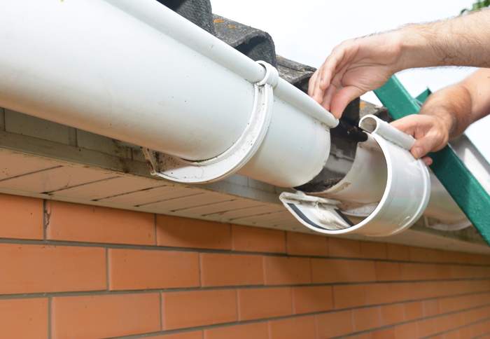 Roof Gutter Installation And Repair A Man On A Ladder Is Replacing A Plastic Rain Gutter Joint Bracket Connecting The Gutters Together